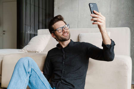 Cheerful unshaven man taking selfie on smartphone while sitting on floor at homeの写真素材