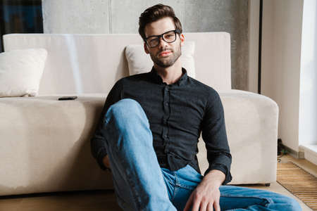 Pleased unshaven man looking at camera while sitting on couch at homeの写真素材