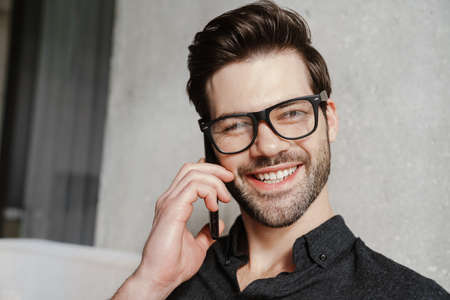 Smiling unshaven man talking on smartphone while sitting on couch at homeの写真素材
