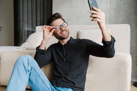 Cheerful unshaven man taking selfie on smartphone while sitting on floor at home