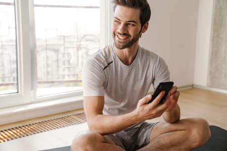 Smiling young sportsman with smartphone sitting on exercise mat at homeの写真素材