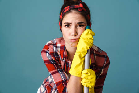 Unhappy tired young woman in gloves posing with mop isolated over blue background