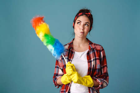 Joyful young woman in gloves smiling while posing with colorful duster isolated over blue backgroundの写真素材