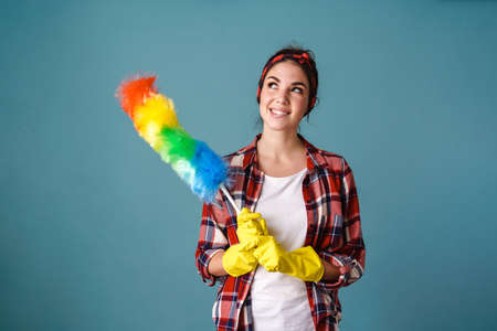 Joyful young woman in gloves smiling while posing with colorful duster isolated over blue backgroundの写真素材
