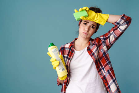 Tired young woman in gloves posing with cleaning product and sponge isolated over blue backgroundの写真素材