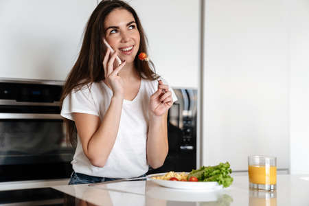 Joyful caucasian woman talking on cellphone while having lunch at home kitchenの写真素材