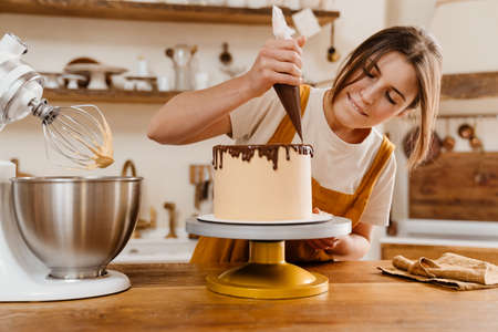 Beautiful focused pastry chef woman making cake with chocolate cream at cozy kitchenの写真素材