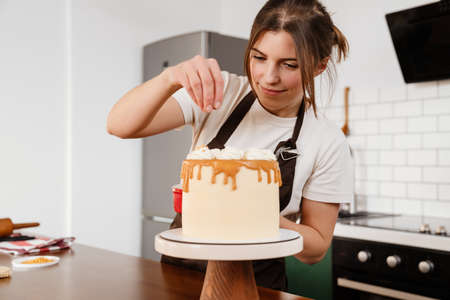 Beautiful pleased baker woman smiling while making cake with cream in kitchen at homeの写真素材