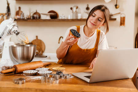Happy pastry chef woman showing tart while making video call on laptop at cozy kitchenの写真素材