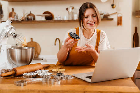 Happy pastry chef woman showing tart while making video call on laptop at cozy kitchenの写真素材