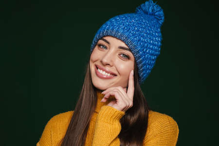 Joyful beautiful girl in knit hat smiling and looking at camera isolated over green backgroundの写真素材