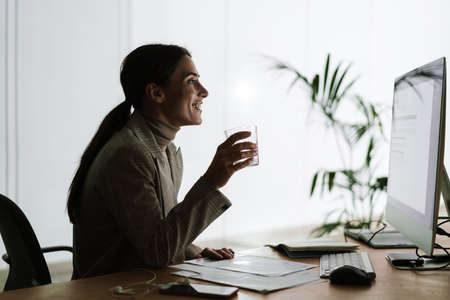 Happy charming woman drinking water while working with computer and papers in officeの写真素材