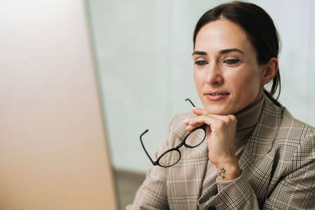 Pleased charming woman holding eyeglasses while working with computer in officeの写真素材