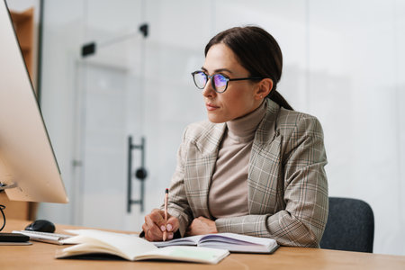 Serious charming woman writing down notes in planner while working at officeの写真素材