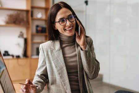 Smiling woman talking on cellphone and working with computer in officeの写真素材