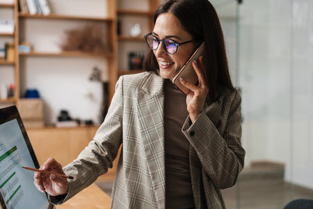 Smiling woman talking on cellphone and working with computer in officeの写真素材