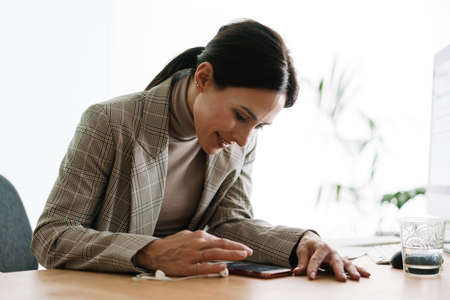 Cheerful charming woman using cellphone while working with computer and documents in officeの写真素材