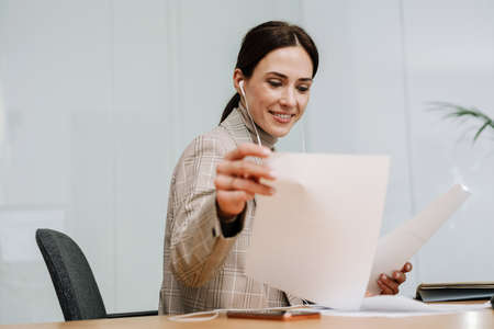 Happy woman in earphones using cellphone while working with papers at office