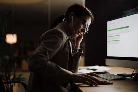 Serious woman in earphones using cellphone while working with computer and papers at office