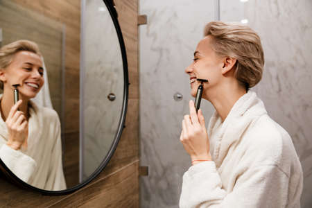Attractive young woman doing a face massage with a cosmetic roller in bathroom in white robe