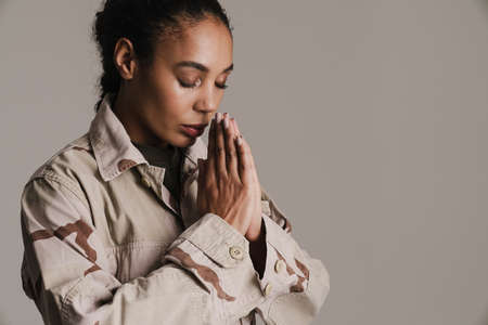 Focused african american soldier woman holding palms together while praying isolated over grey wall