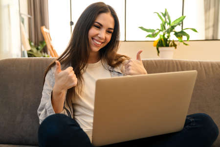 Happy young woman showing thumbs up while using laptop indoors