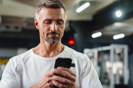 Pleased grey adult sportsman using cellphone and working out in fitness roomの写真素材