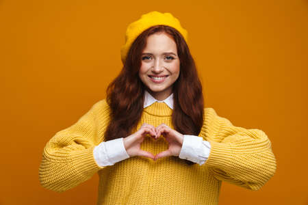 Happy young woman with long red hair in sweater and beret standing over yellow wall background, showing love gestureの写真素材