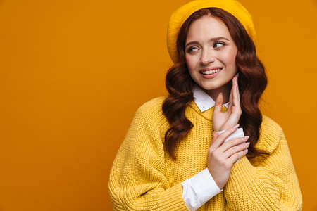 Happy young woman with long red hair in sweater and beret standing over yellow wall background, lookng aside at copy spaceの写真素材