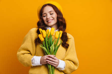 Smiling young woman with long red hair in sweater and beret standing holding tulips bouquet over yellow wall backgroundの写真素材
