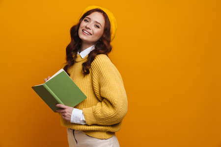 Happy young woman with long red hair in sweater and beret standing over yellow wall background, holding paper bookの写真素材
