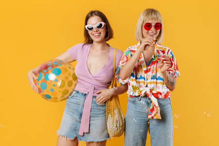 Portrait of two cheerful lovely girlfriends wearing summer clothes standing isolated over yellow background, holding beach ball, drinking beverage, blow soap bubblesの写真素材
