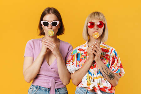 Portrait of two cheerful lovely girlfriends wearing summer clothes standing isolated over yellow...