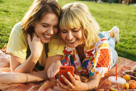 Image of joyful beautiful two women laughing and using cellphone while have picnic on grass in summer parkの写真素材