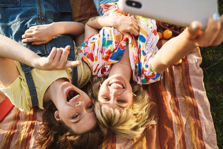 Image of joyful two women taking selfie and pointing at cellphone while lying on blanket in summer parkの写真素材