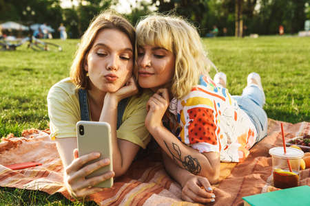 Image of joyful beautiful two women taking selfie on cellphone while have picnic on grass in summer parkの写真素材