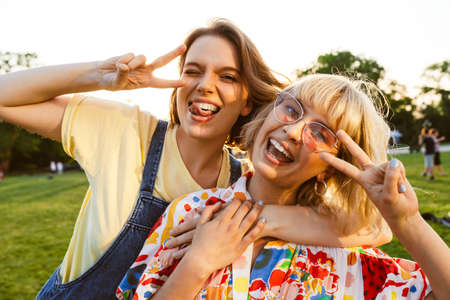 Image of laughing two women gesturing peace sign and hugging while walking in summer park