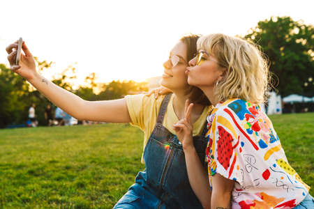 Image of funny two women gesturing peace sign and taking selfie on smartphone while resting in summer parkの写真素材
