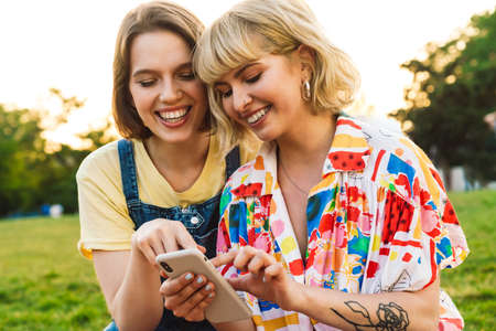 Image of cheerful beautiful two women laughing and sing mobile phone while resting on grass in summer parkの写真素材