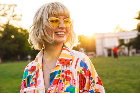 Image of happy young woman in sunglasses smiling and drinking soda while resting in summer park