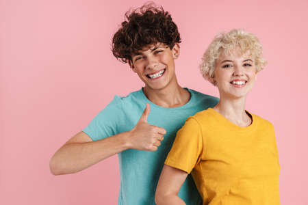 Happy young couple of teenagers standing isolated over pink background, wearing t-shirts, giving...