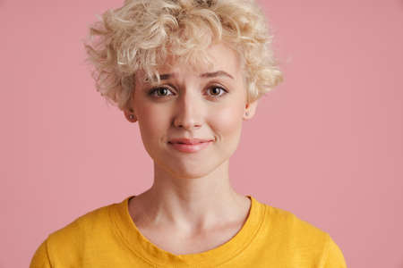 Portrait of a young confused blonde girl with curly blonde hair standing isolated over pink...