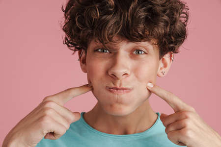 Young happy teenager with curly hair blowing his cheeks isolated over pink background