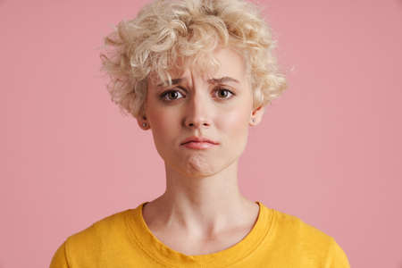 Portrait of a young upset blonde girl with curly blonde hair standing isolated over pink backgroundの写真素材