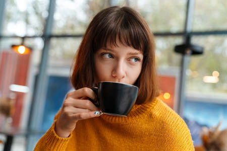 Beautiful redhead girl drinking coffee while sitting in cafe indoorsの写真素材