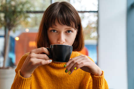 Beautiful redhead girl drinking coffee while sitting in cafe indoorsの写真素材