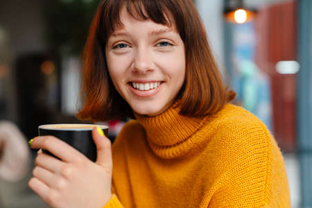 Happy redhead girl smiling and drinking coffee while sitting in cafe indoors