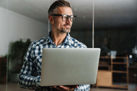 Pleased handsome man working with laptop while standing in office