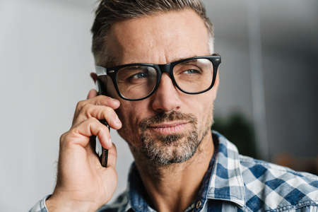 Focused white-haired man in eyeglasses talking on cellphone in officeの写真素材