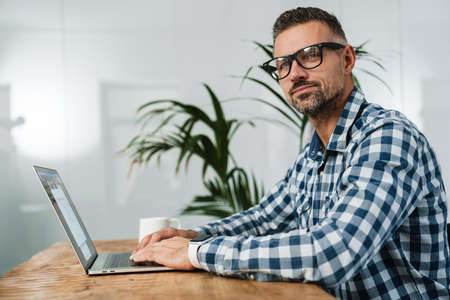 Focused unshaven man in eyeglasses working with laptop in officeの写真素材